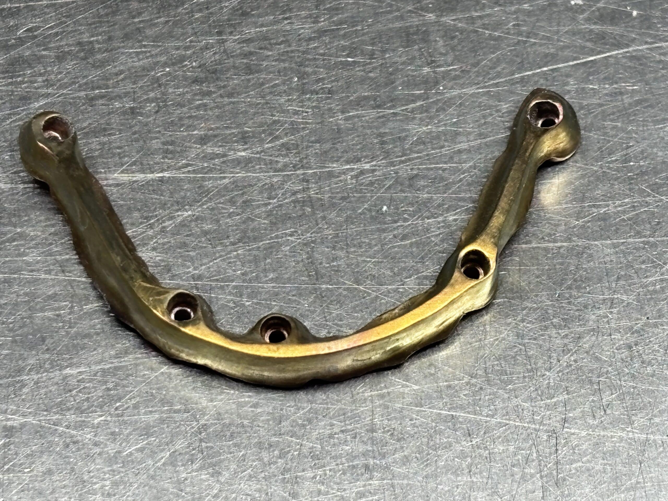 Armature dentaire métallique dorée avec perforations pour implants, posée sur une surface en acier brossé.